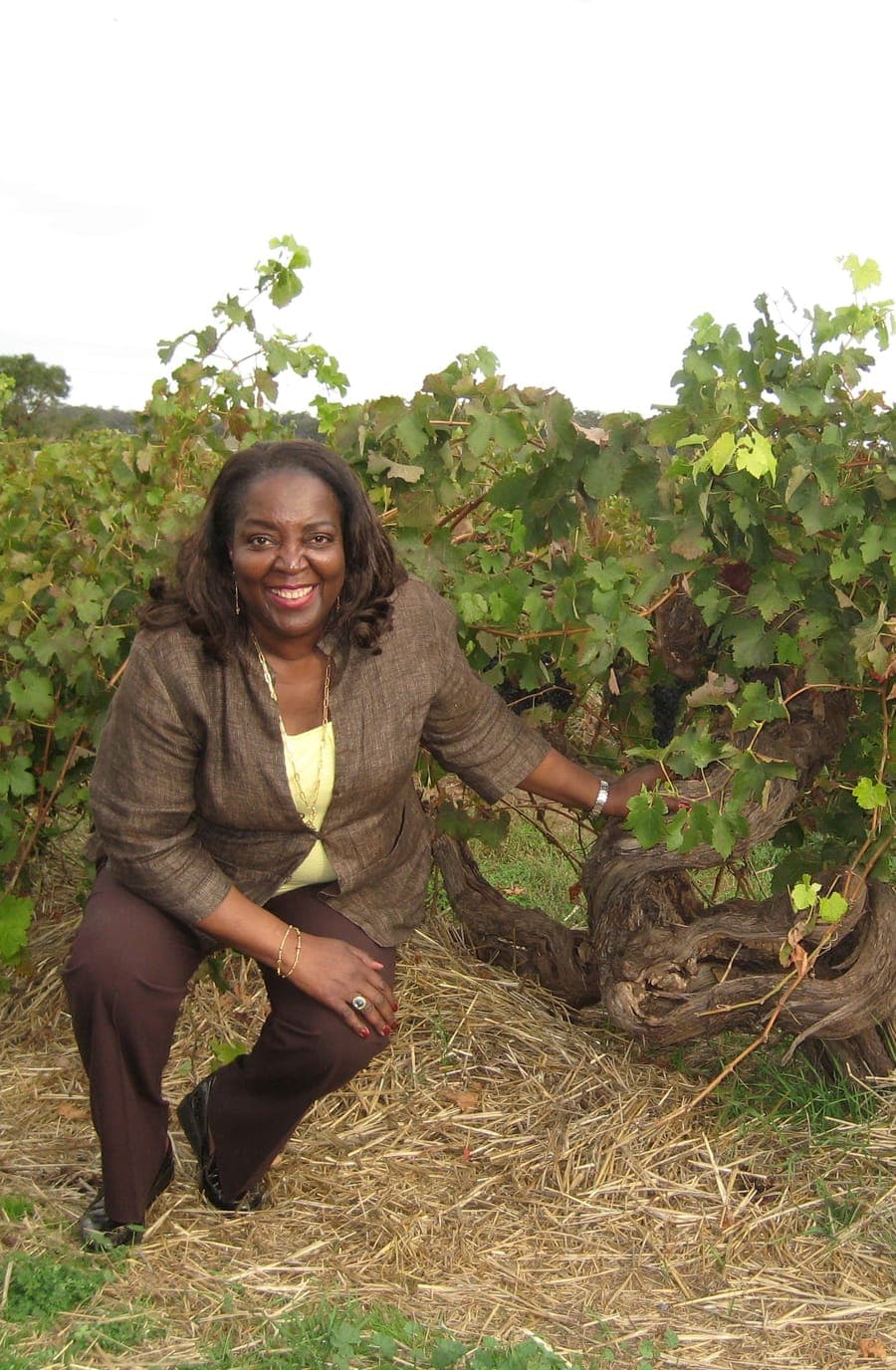 Sandra E. Taylor crouching beside an old gnarled grapevine, one hand resting on its trunk